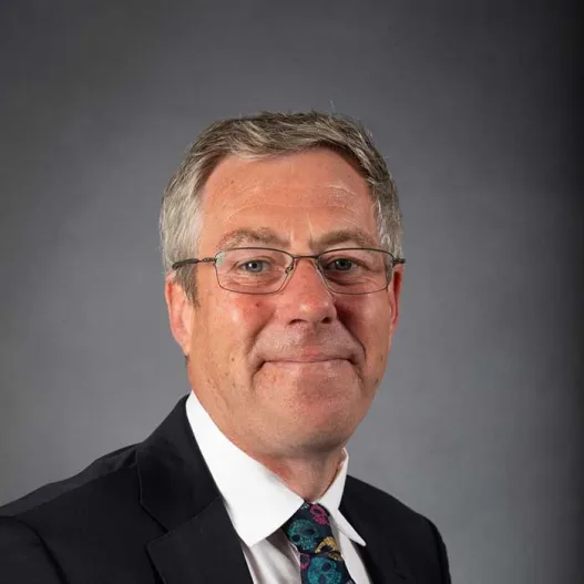 A middle-aged man with glasses wearing a dark suit, white shirt, and a colourful patterned tie, posing against a plain grey background.