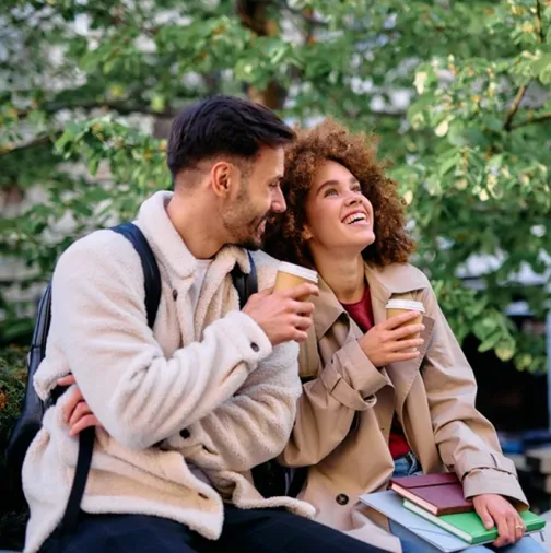 Two people sit outdoors, holding coffee cups and smiling, with books and greenery in the background.