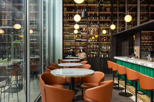 Modern bar with orange chairs, marble tables, globe lights, and a wall of shelves filled with bottles behind the counter.