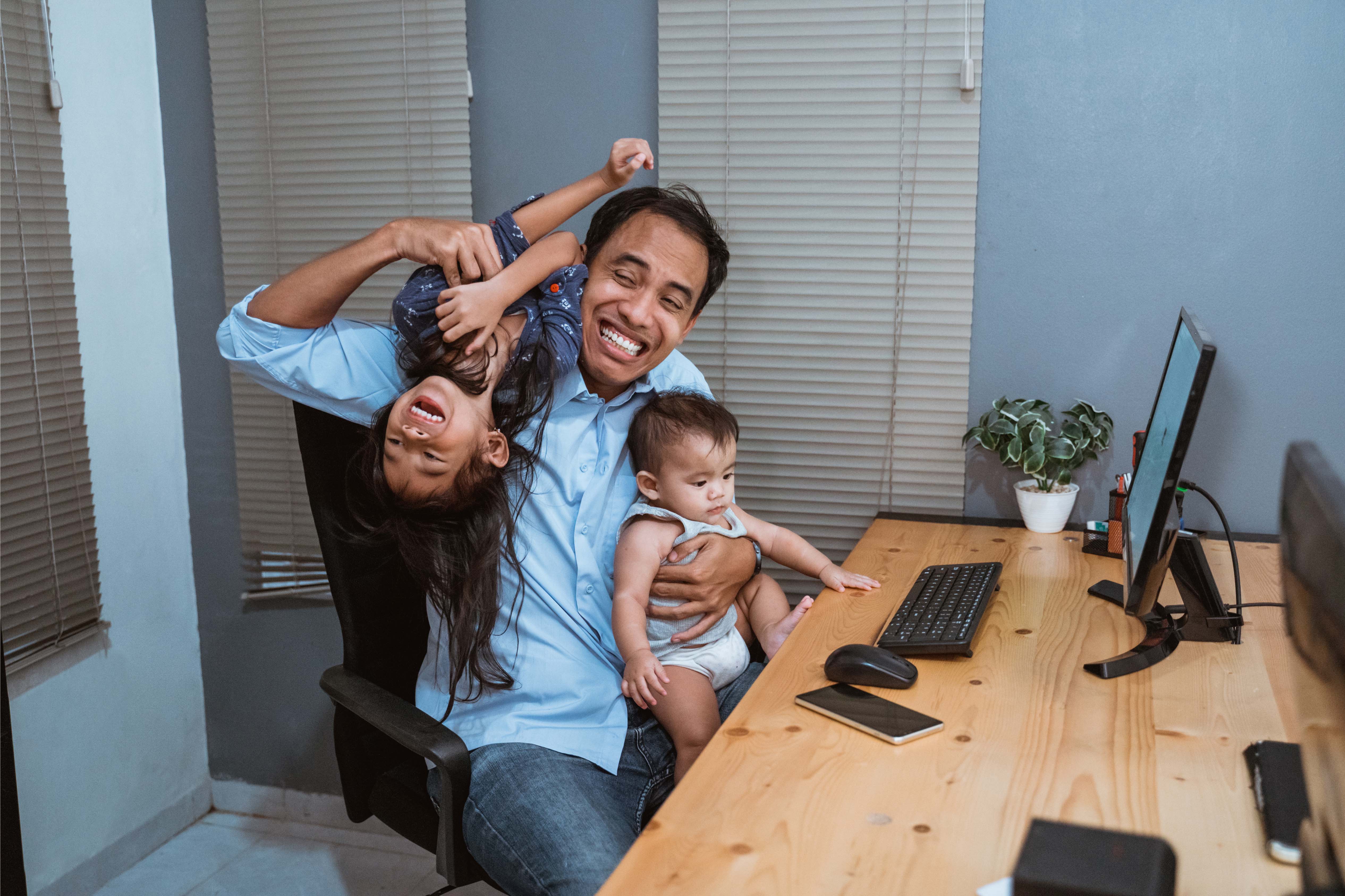 Indonesian father working at home on a computer with his daughter on his back and baby on his lap, representing work-life balance and family-focused flexible benefits