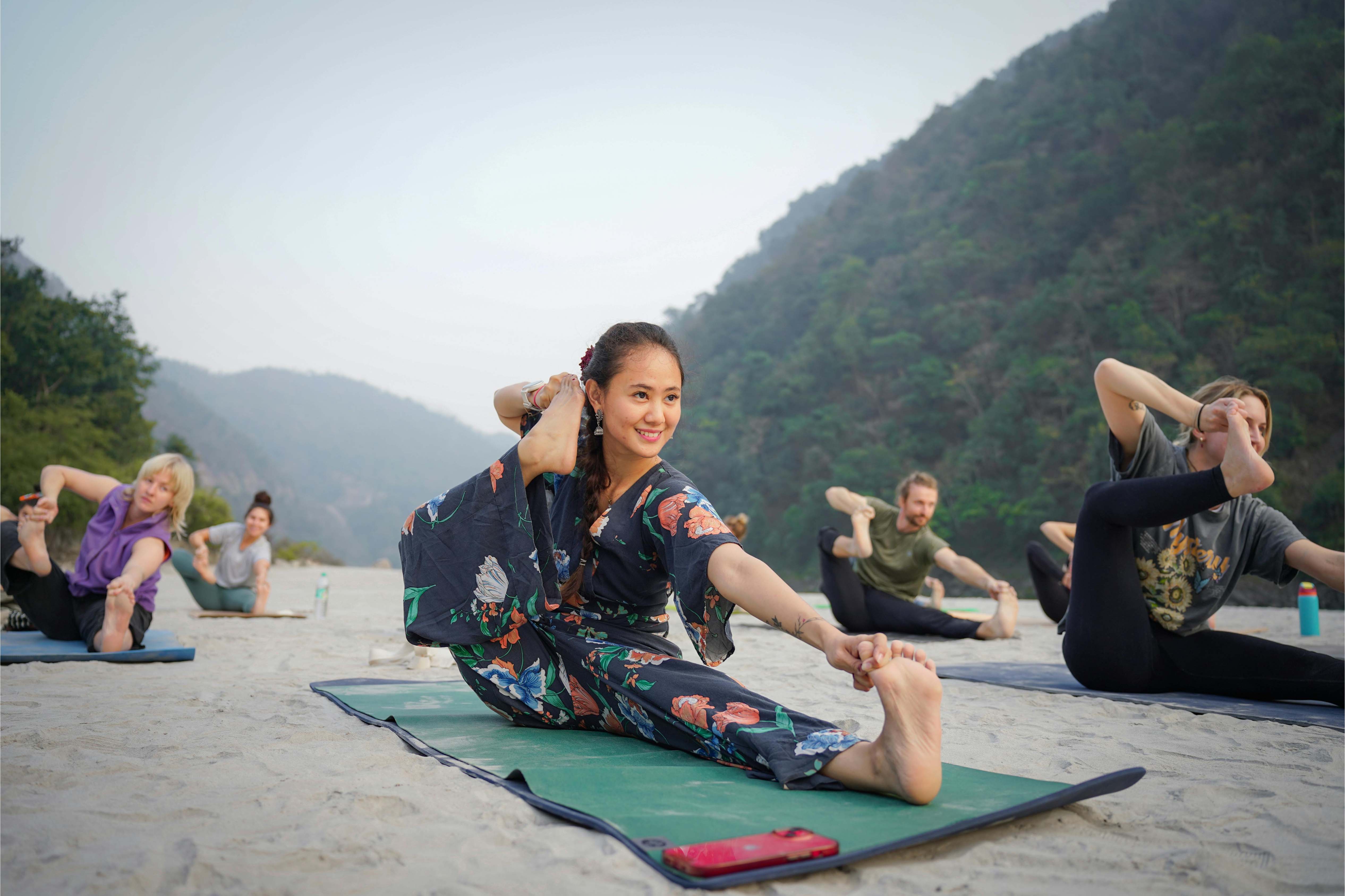 A woman in floral activewear leads a yoga group on a sandy beach with lush green mountains in the background, promoting wellness and mindfulness.
