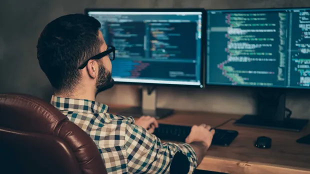 A bearded man in glasses sits in a leather chair at a wooden desk, coding across two monitors