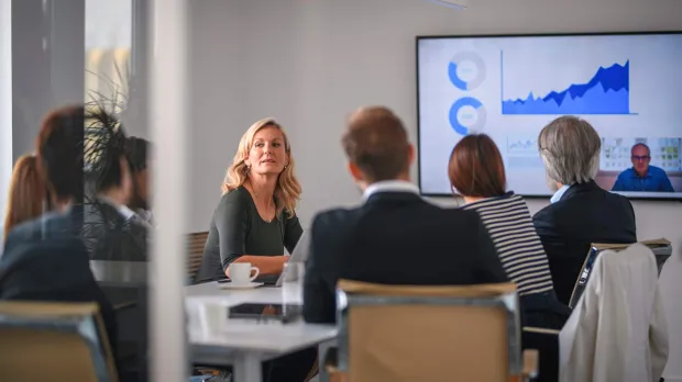  blonde business woman speaks to a colleague during a board meeting, where everyone around the table sits looking at a presentation on a large screen
