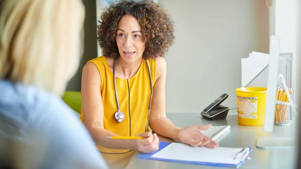 A female doctor talks a patient through her medical chart. Both women sit at the doctor's desk, and the doctor is wearing a stethoscope around her neck
