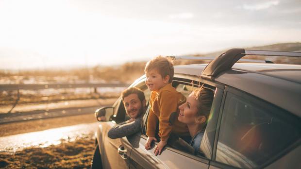 A young family take a day trip to the beach. They sit in their car looking out of the windows at the sea. The dad sits in the driver's seat, while his wife and son sit in the back seat. The little boy leans out of the window to get a better view.
