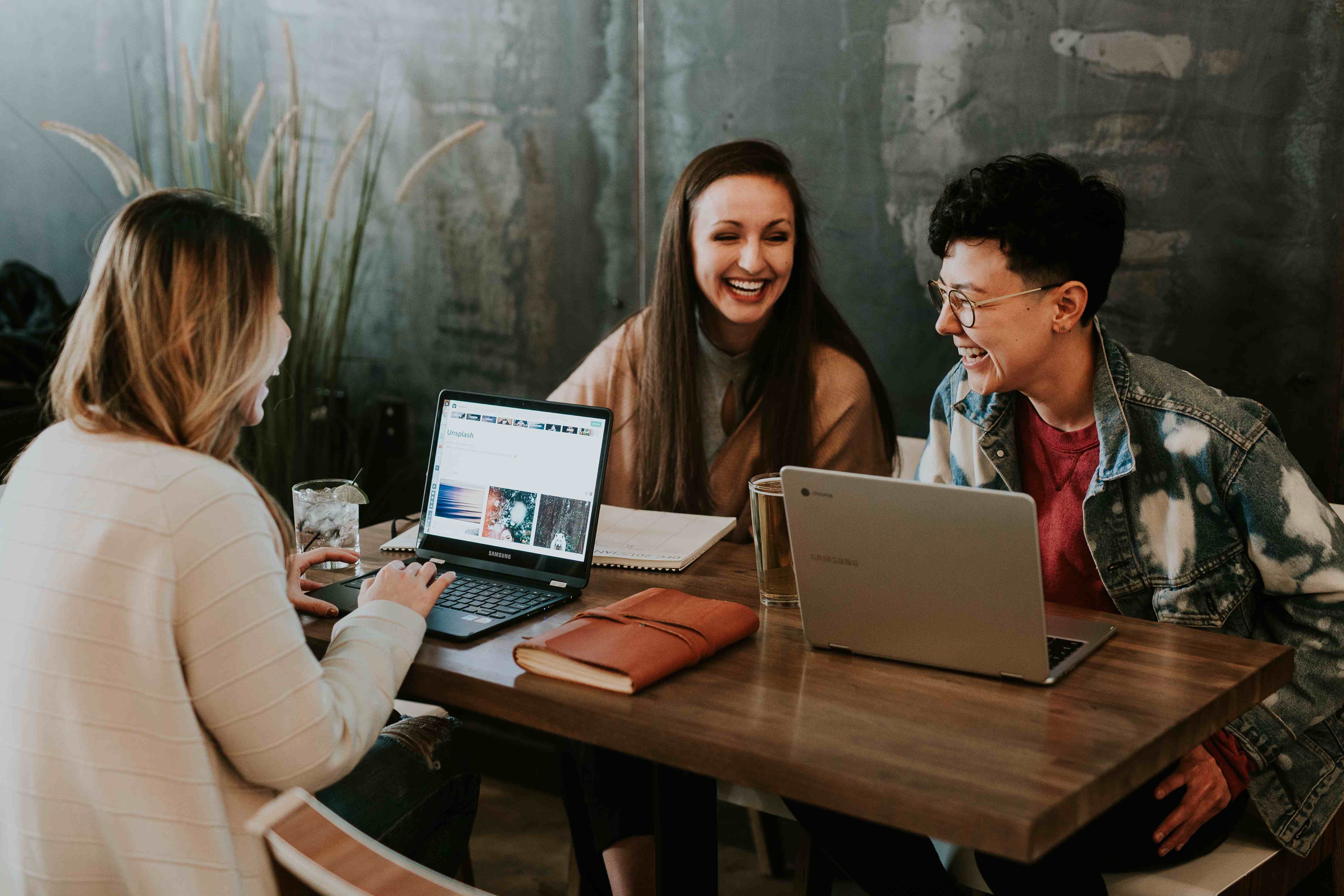Three colleagues smiling and working together on laptops at a wooden table in a relaxed, modern workspace