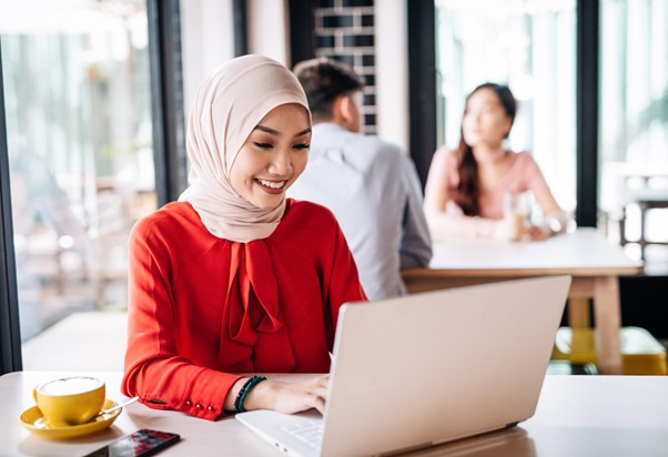 Smiling woman in a light hijab and red blouse typing on a laptop at a café table with a cup of coffee nearby