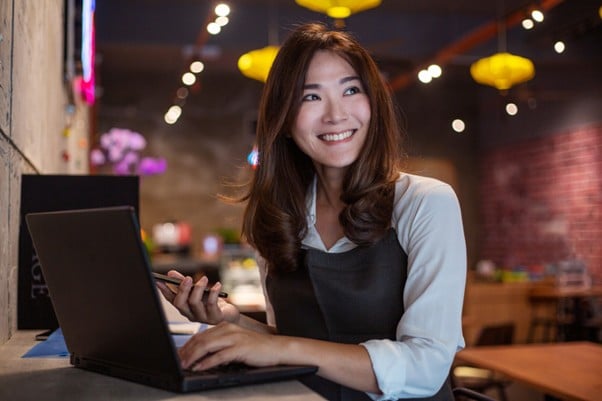 Smiling woman using a laptop and holding a phone while working from a dimly lit café