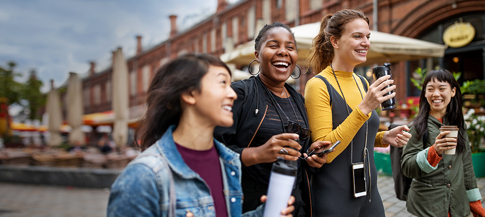 A diverse group of young women laughing and holding coffee cups while walking outdoors in a city setting