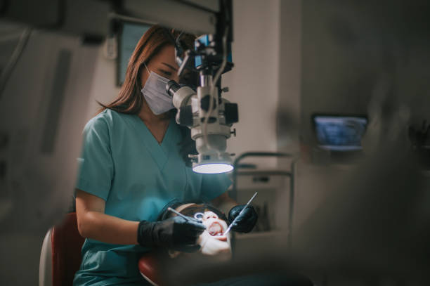 Female dentist wearing scrubs and a surgical mask using a dental microscope to treat a patient in a clinical setting