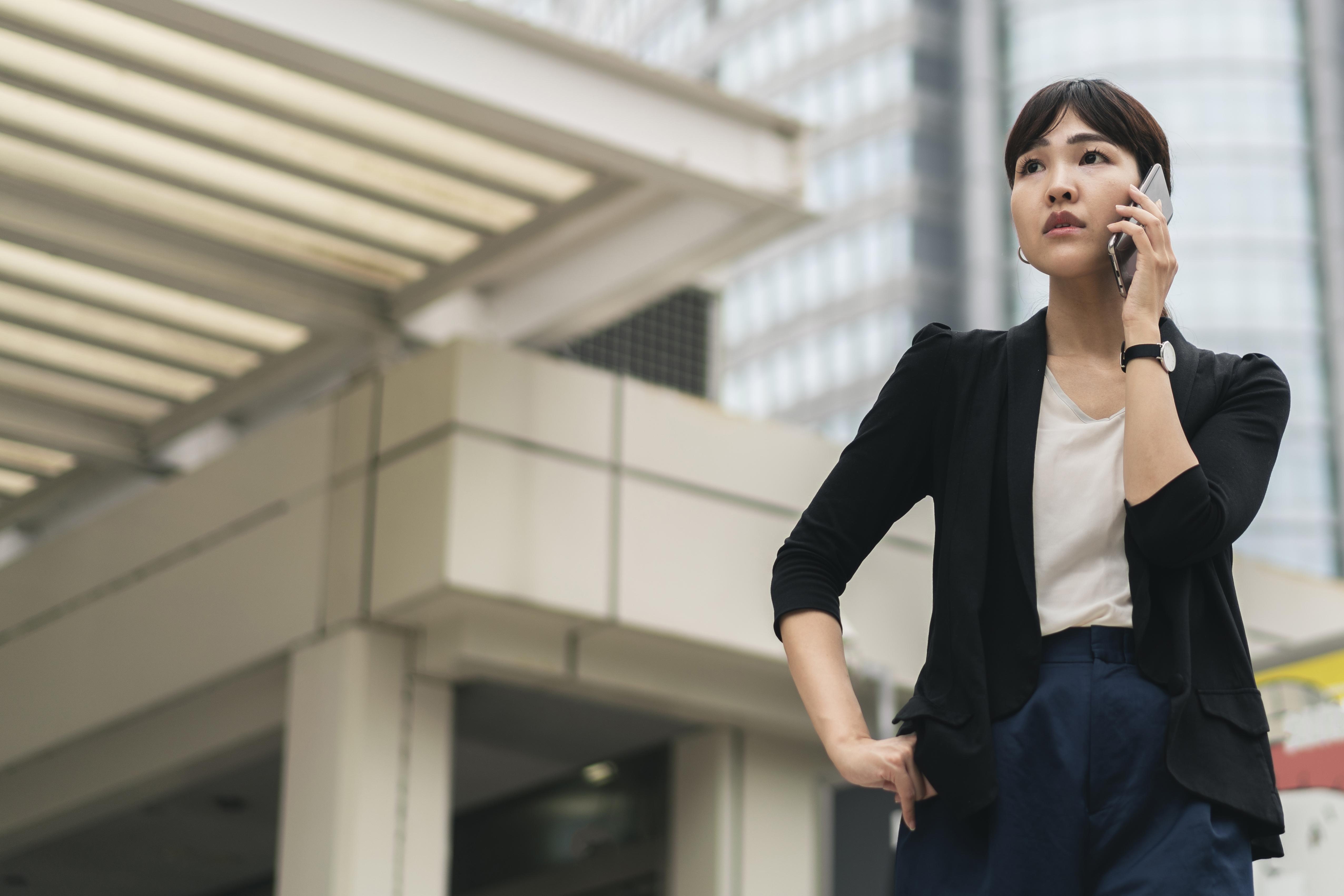 Asian businesswoman in smart attire speaking on the phone while standing outside a commercial building in an urban setting