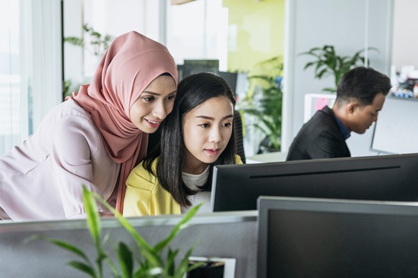 Two professional women, one wearing a hijab, looking at a computer screen together in a shared office space
