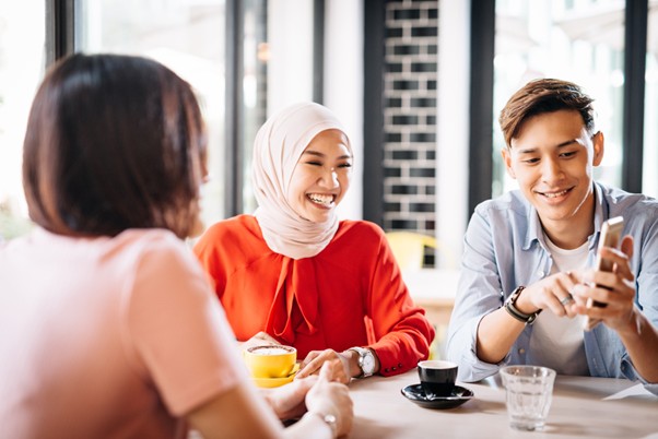 Group of young adults, including a woman in a hijab, smiling and chatting around a café table with drinks and a smartphone