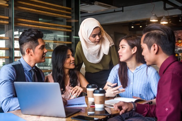 Team of professionals, including a woman in hijab, having a strategy conversation in a modern café-style meeting area with coffee