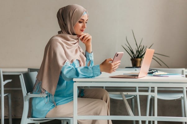 Malaysian woman in a hijab working at a laptop and holding her smartphone in a contemporary workspace