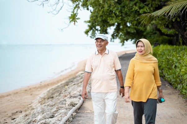 Smiling older Malaysian couple walking hand-in-hand along a coastal path, representing health and wellbeing in retirement