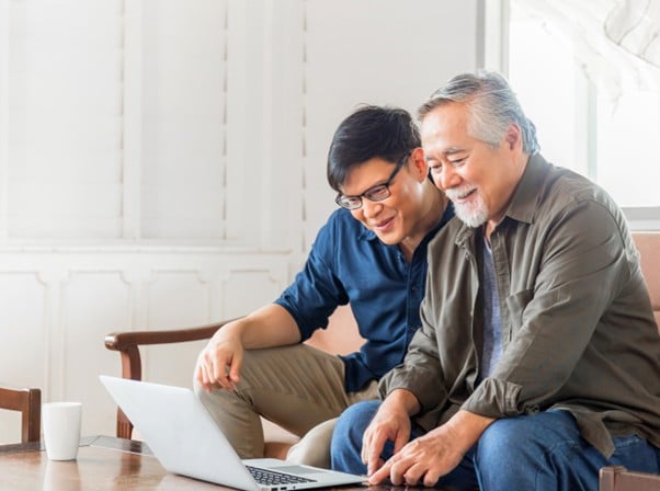 A younger man and his elderly father looking at a laptop and smiling, symbolising intergenerational financial planning.