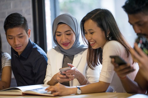Two women, one in a hijab, smiling and looking at a mobile phone with colleagues in a casual work environment