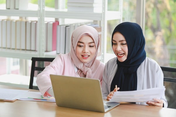 Two Malaysian Muslim women in hijabs working together on a laptop in a bright office