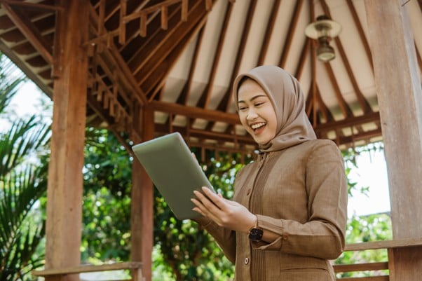 Malaysian woman in a hijab using a tablet while smiling in a wooden outdoor workspace