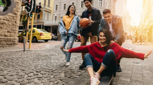 A group of young Malaysian adults having fun together on a city street, laughing and skateboarding, with a bright urban backdrop