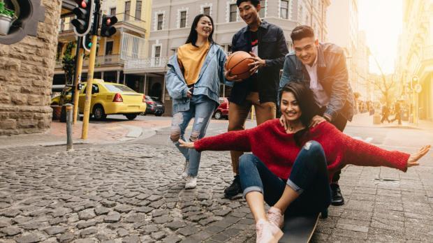 A group of young Malaysian adults having fun together on a city street, laughing and skateboarding, with a bright urban backdrop