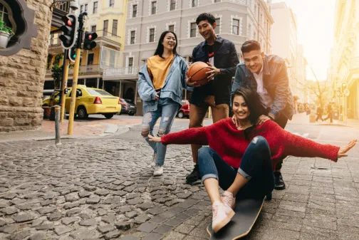 A group of young Malaysian adults having fun together on a city street, laughing and skateboarding, with a bright urban backdrop