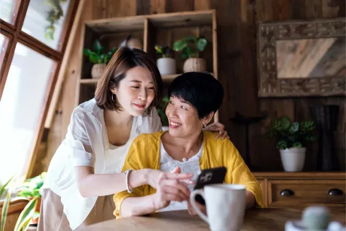 Affectionate Asian senior mother and daughter using smartphone together at home, smiling joyfully, enjoying mother and daughter bonding time. Multi-generation family and technology