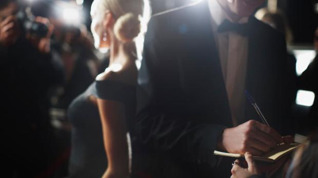 A man in a tuxedo signs an autograph book while a blonde woman in a ballgown is photographed by paparazzi behind him