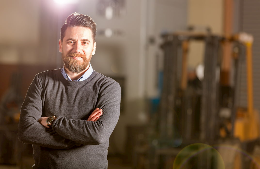 A man in a factory wearing a shirt and jumper smiles at the camera with his arms crossed