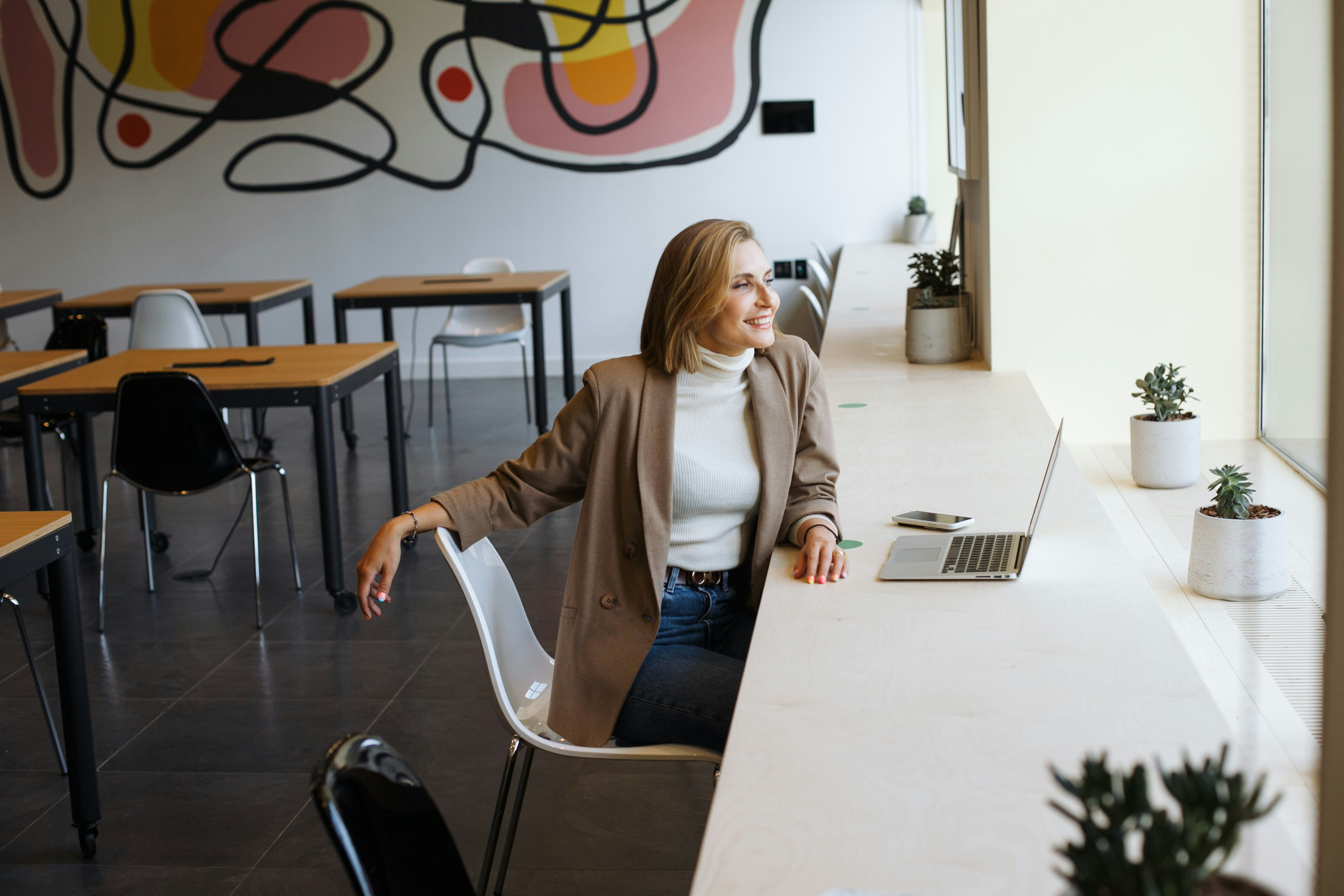 A smiling woman in a beige blazer sits at a long desk in a modern, brightly lit office space, looking out the window. A laptop and phone rest on the desk, with small potted plants placed along the surface.