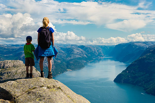 A woman and child stand on a cliff edge overlooking a scenic Norwegian fjord, with backpacks and hiking gear, enjoying the panoramic view of mountains and water.