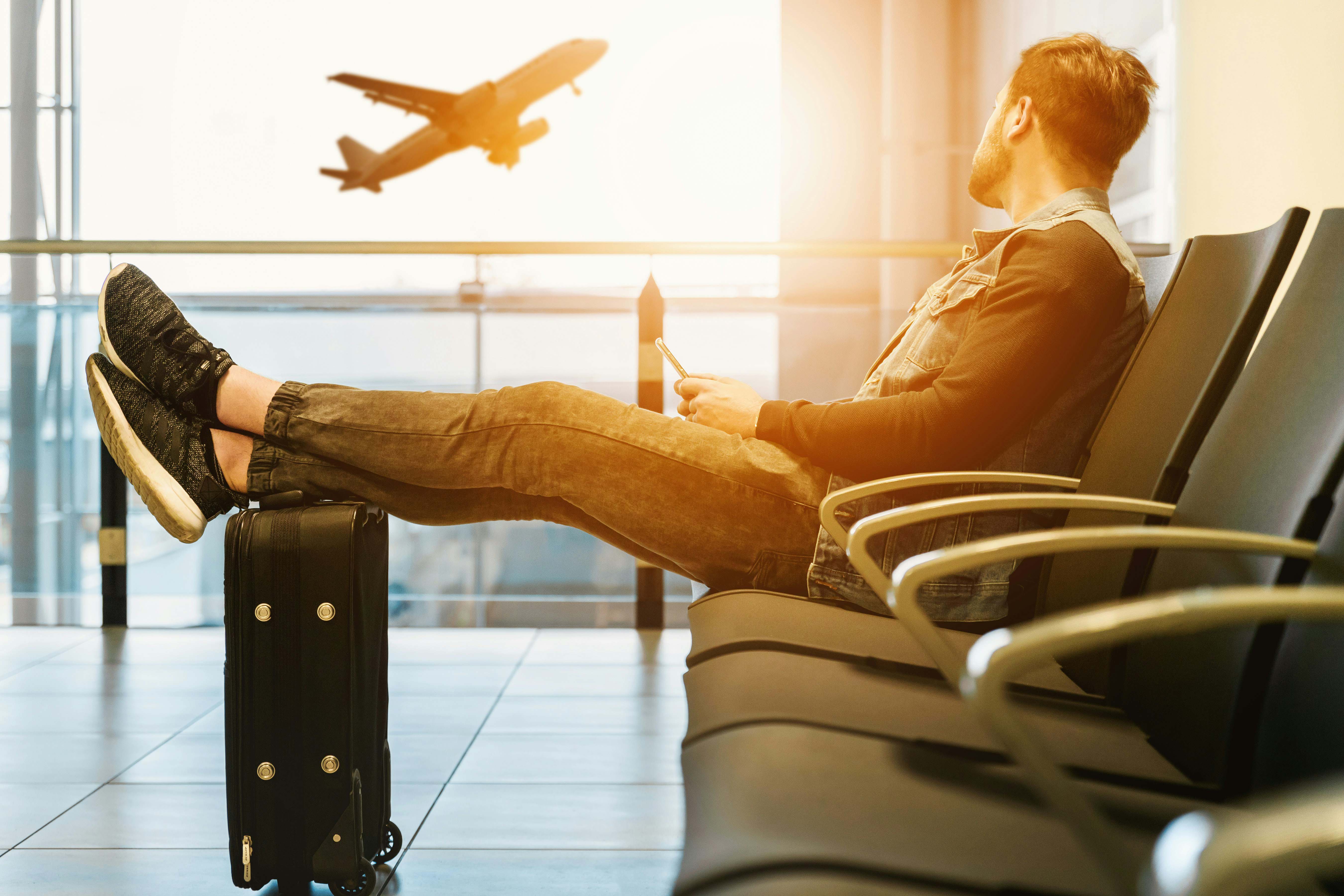 A man sits in an airport lounge with his legs resting on a suitcase, looking out at a plane taking off through the window during sunset.