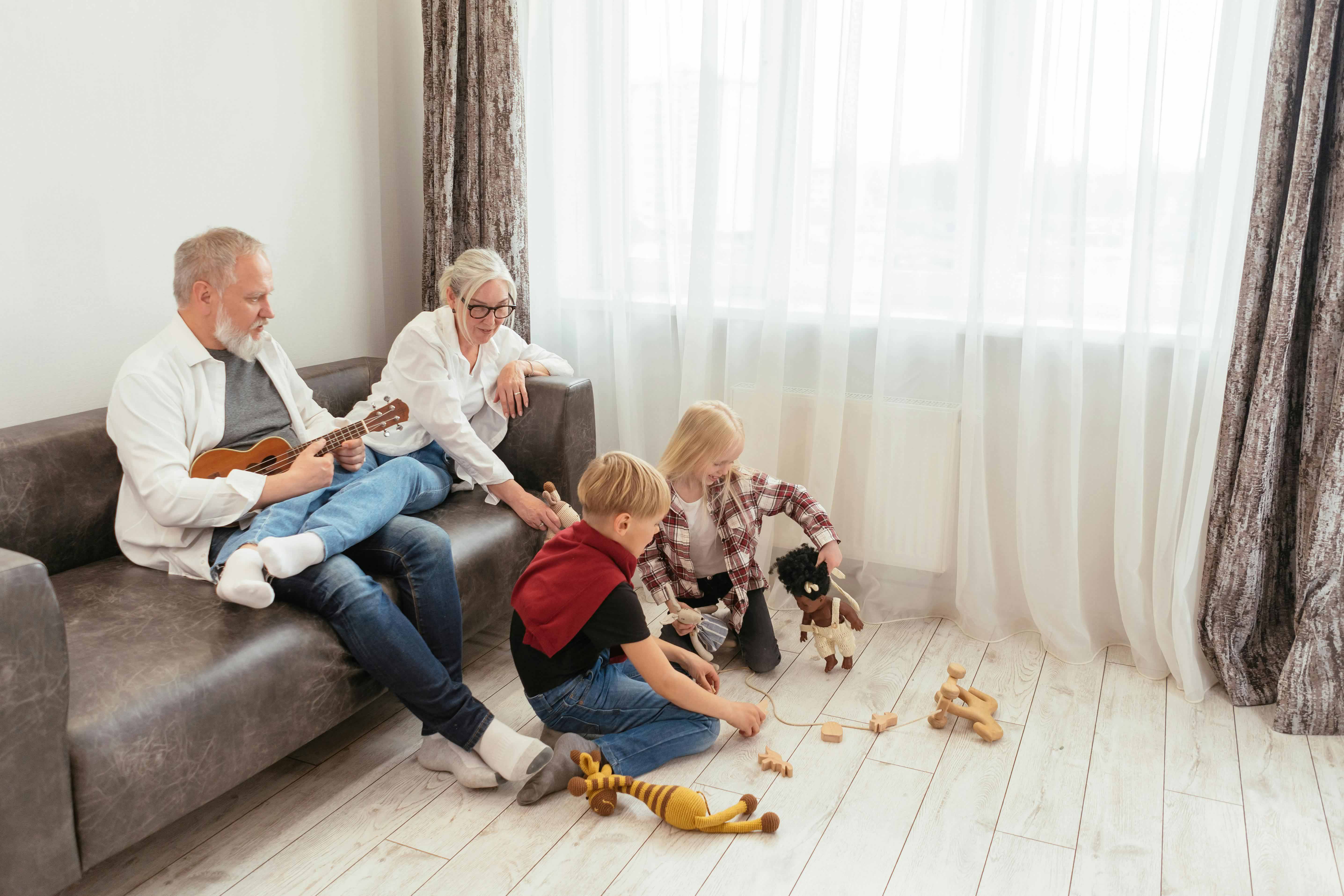 Two grandparents sit on a sofa, watching and playing with their two young grandchildren who are sitting on the floor with wooden toys and dolls in a bright living room.