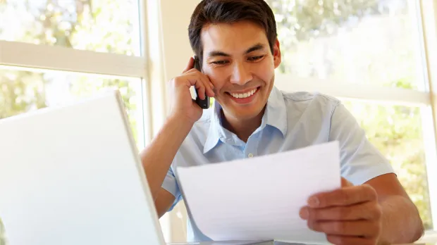 Man talking on the phone and checking paperwork at his desk Man reviewing documents while speaking on the phone and working on a laptop, representing support and easy communication for SMEs.