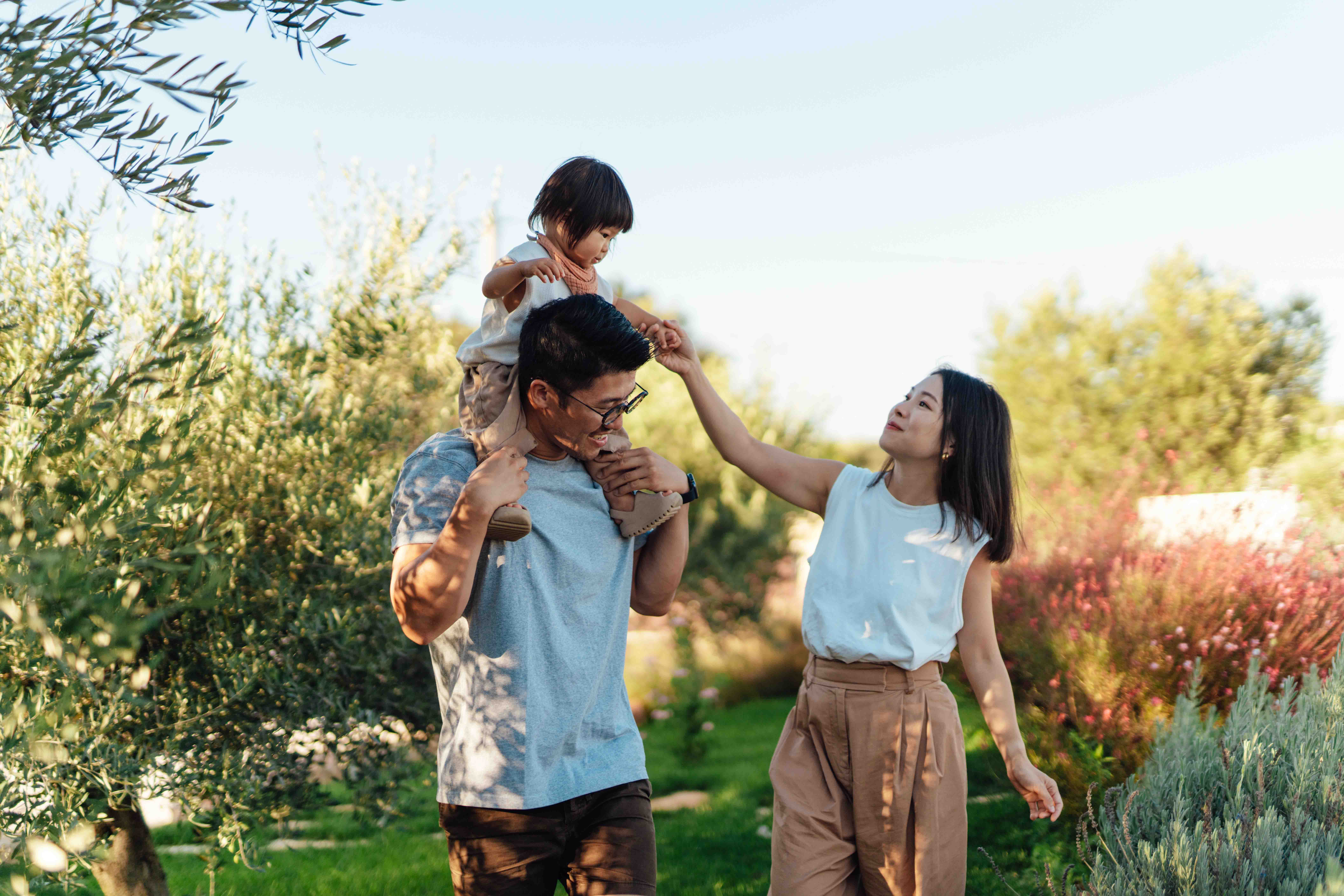 Young family enjoying time together outdoors, with a child on their father’s shoulders and mother playfully reaching out, surrounded by greenery.