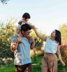 Young family enjoying time together outdoors, with a child on their father’s shoulders and mother playfully reaching out, surrounded by greenery.