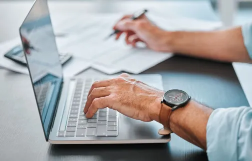 man typing on a laptop and writing on a notepad