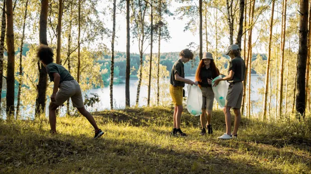 Un grupo de adolescentes limpia un bosque junto a un lago, recogiendo basura en bolsas de plástico.