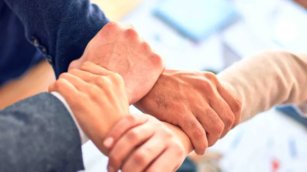 Primer plano de manos diversas juntándose sobre una mesa en un entorno de oficina, simbolizando trabajo en equipo y cooperación.