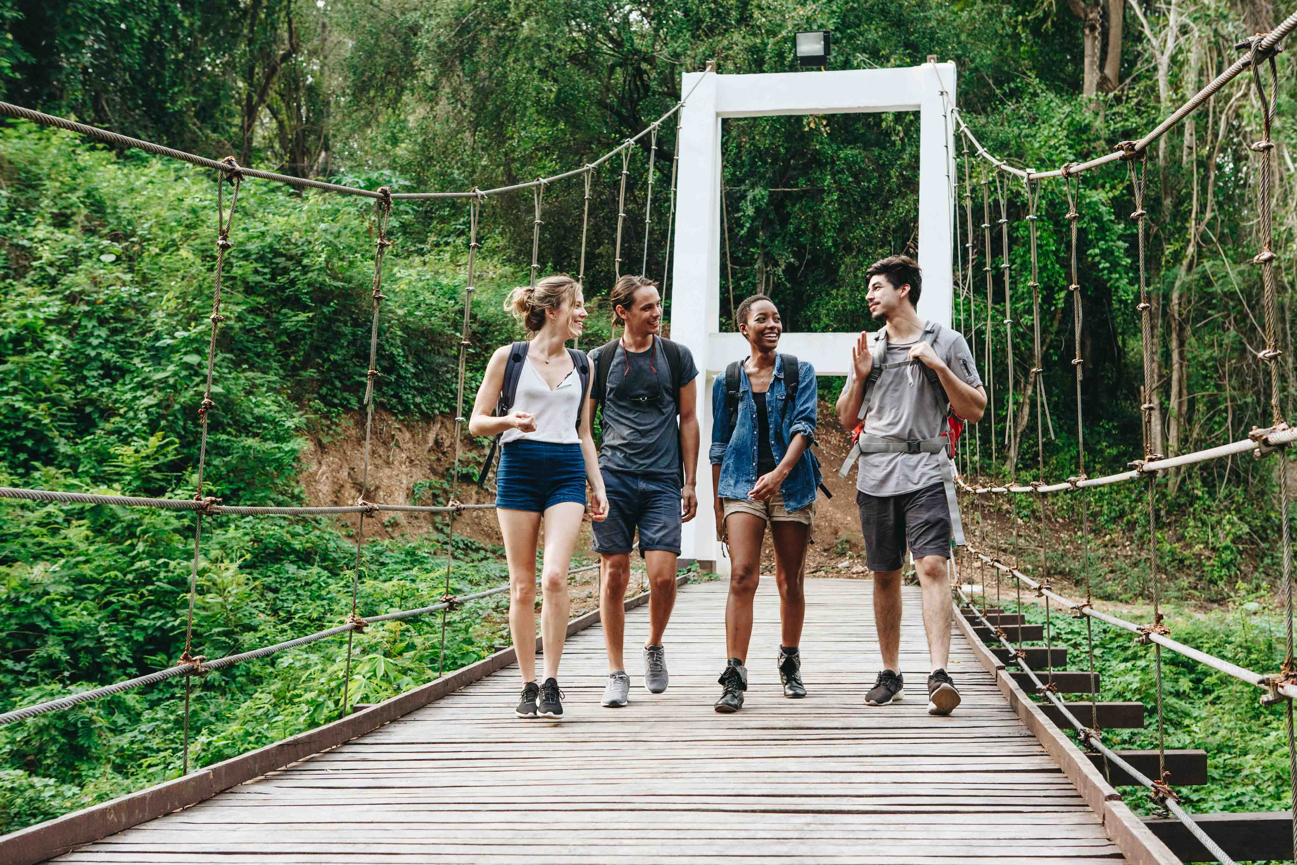 Four young professionals walking across a suspension bridge in a lush green forest, wearing backpacks and talking.