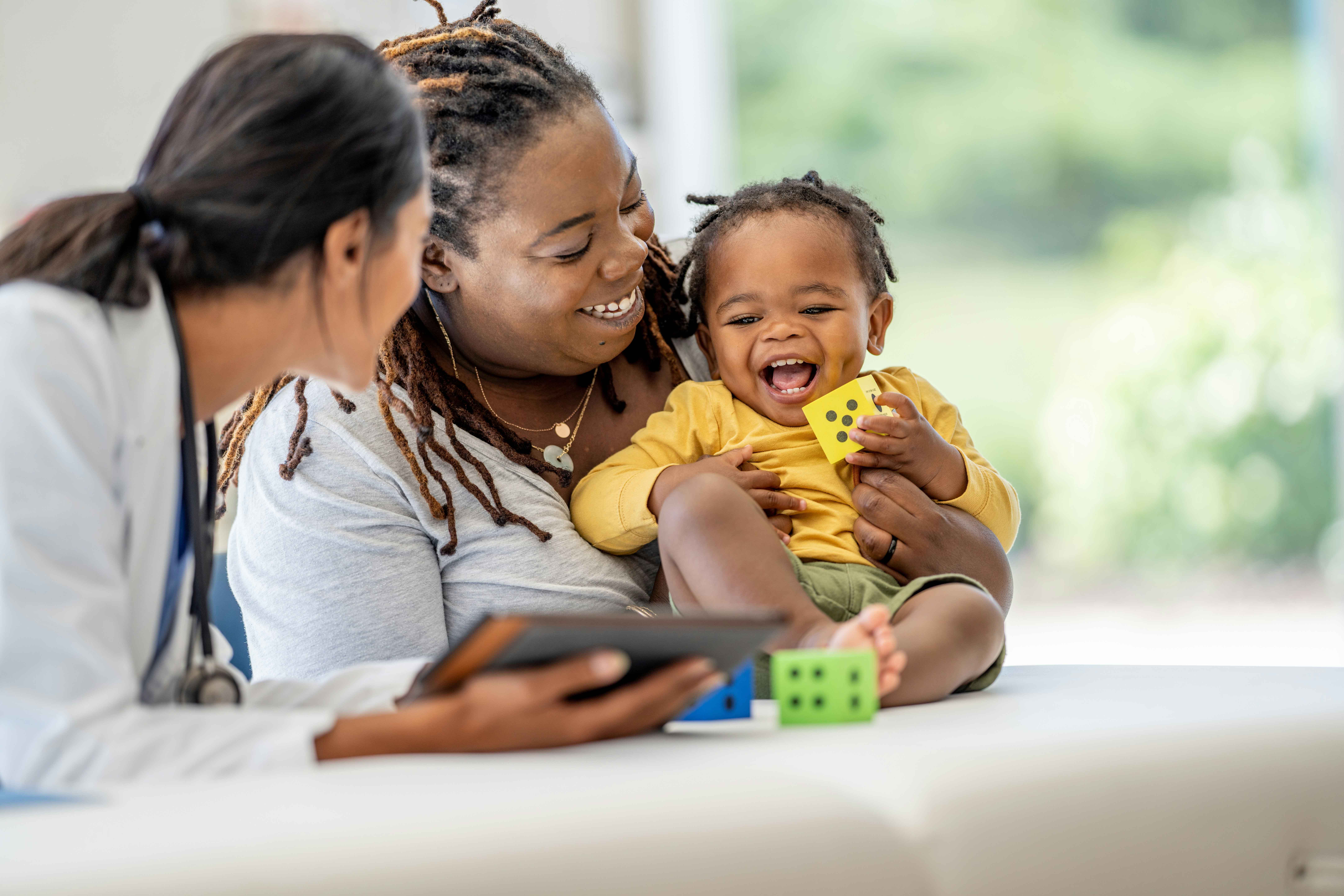 Mother holding a smiling toddler while speaking with a healthcare professional during a clinic visit.