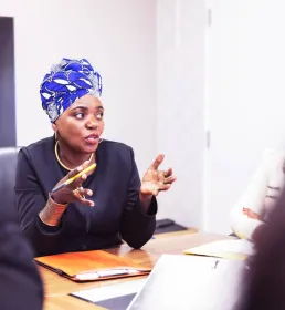 Corporate business woman sits at a board table with her colleagues. She is speaking and gesturing with her hands.