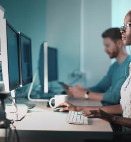A female IT technician sits at her desk next to a male colleague. They are both working on their respective computers. She is wearing headphones and looking at her two monitors.