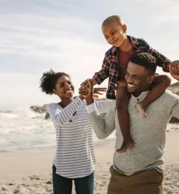 A young family of three walk along the beach. The father carries his happy son on his shoulders and holds his hands. The mother walks next to them and reaches towards her son. All three of them have big smiles