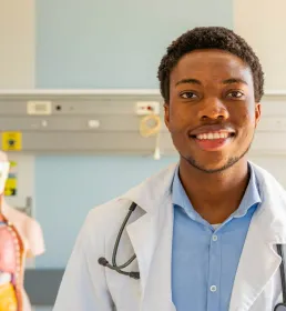 A young doctor in a white coat and blue shirt stands on a hospital ward. He is wearing a stethoscope and standing in front of an anatomical model.