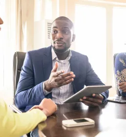 A professional man in a tailored suit sits at a table with two business women. He is holding a tablet computer and speaks directly to one of the women.