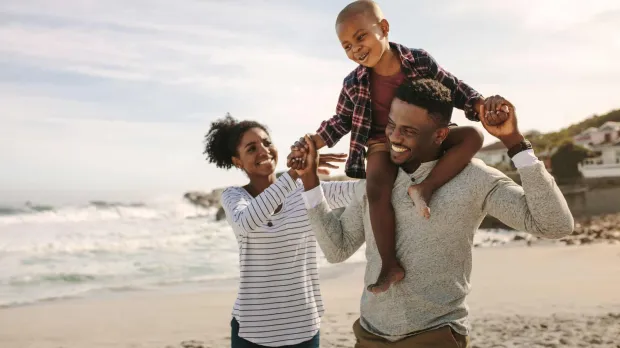 Employee Benefits - family on the beach A young family of three walk along the beach. The father carries his happy son on his shoulders and holds his hands. The mother walks next to them and reaches towards her son. All three of them have big smiles