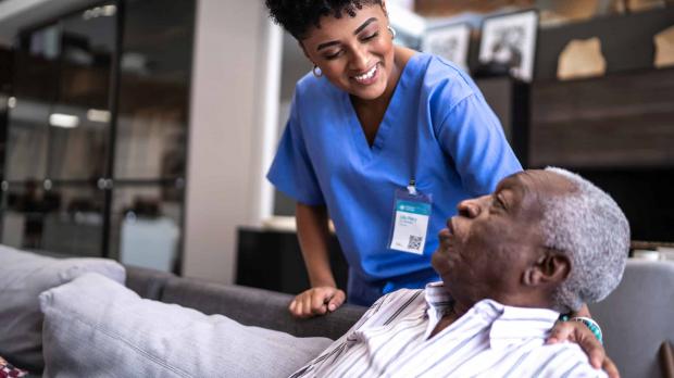 A friendly female nurse in blue scrubs leans over the back of a sofa to speak with her older male patient