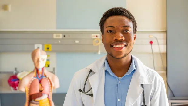 Medical Malpractice - smiling doctor A young doctor in a white coat and blue shirt stands on a hospital ward. He is wearing a stethoscope and standing in front of an anatomical model.